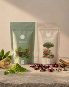 Two stand-up pouches of herbal tea or matcha-style products on a stone surface: a sage green pouch labeled Midori with green powder in front, and a cream pouch labeled Socotra with botanical artwork. Decorative leaves, dried red flowers, berries, and spices are arranged around them against a warm beige background.