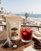 A stand-up pouch of Socotra tea sits on a wooden table beside a glass of iced red beverage, with a beach and ocean visible in the background. A tea bag and sunglasses are also on the table.